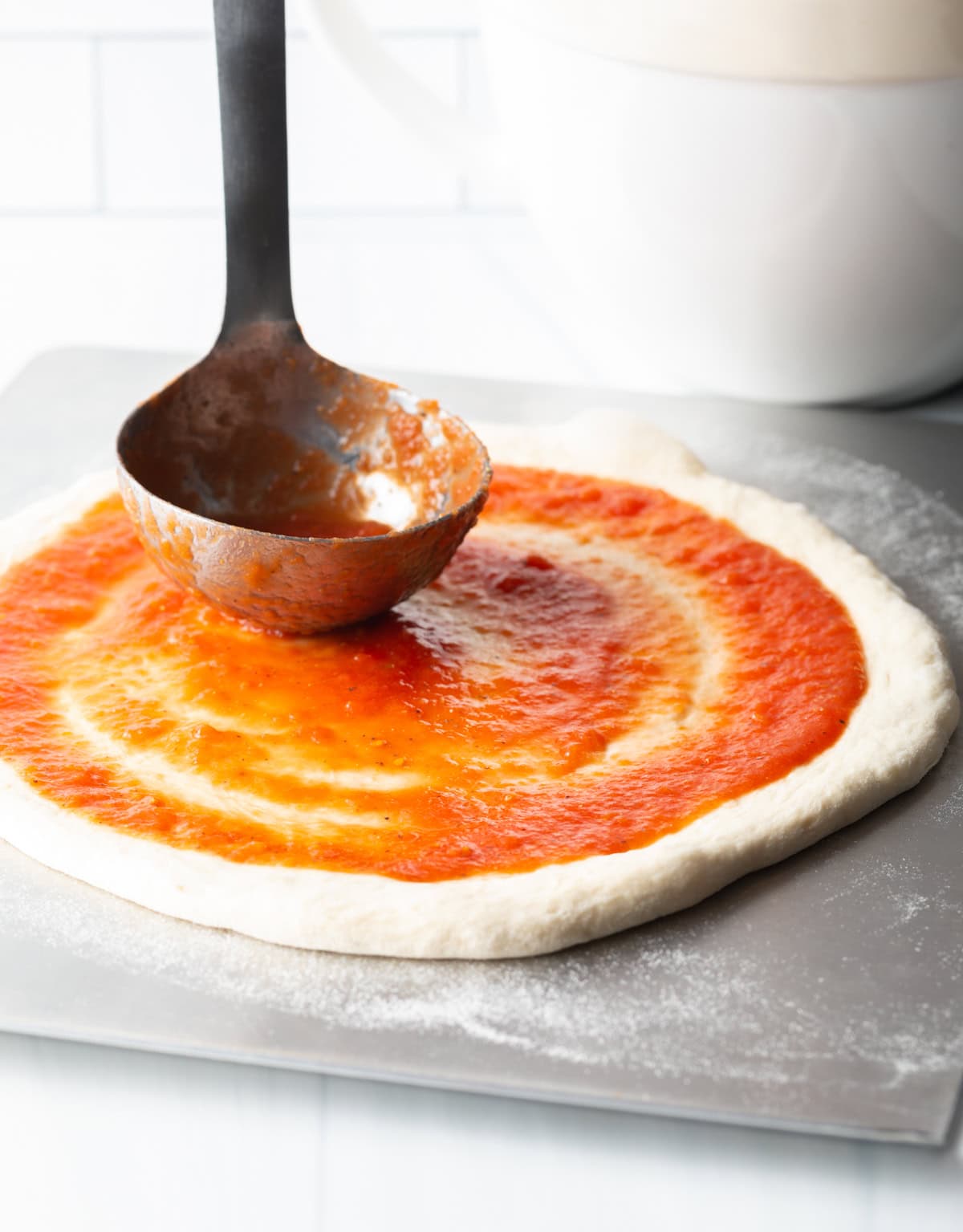 Using a metal ladle to smear tomato sauce across homemade pizza dough.