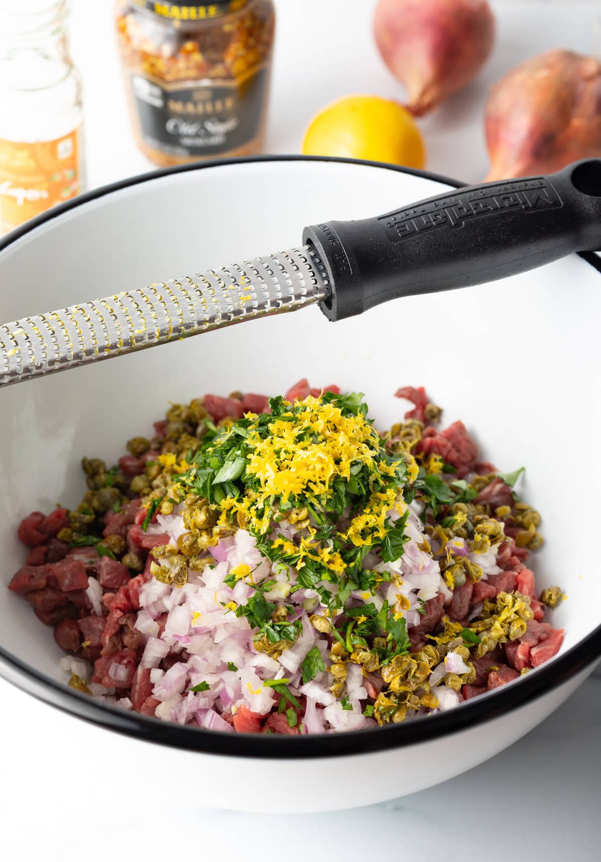 Bowl of measured and chopped steak tartare ingredients, with a microplane resting on the edge of the bowl.