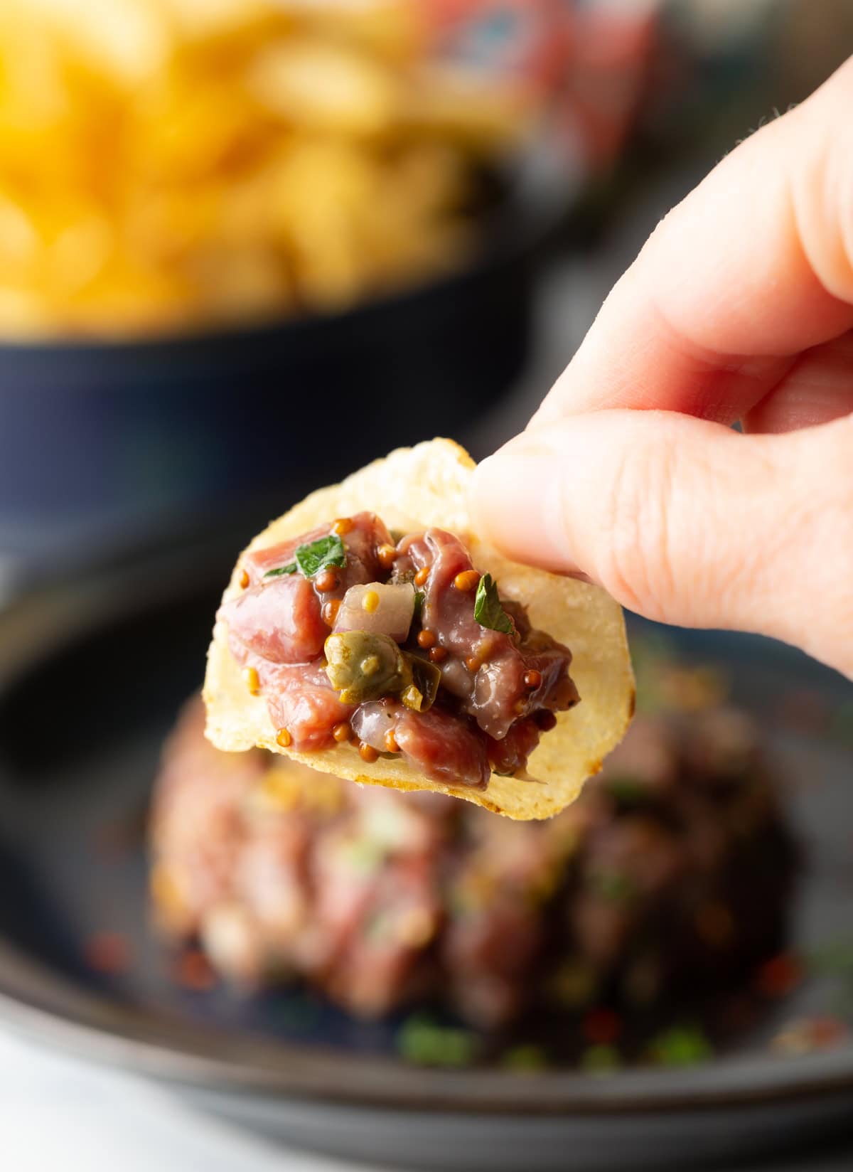 Scoop of beef tartare on a chip being held between a thumb and forefinger.