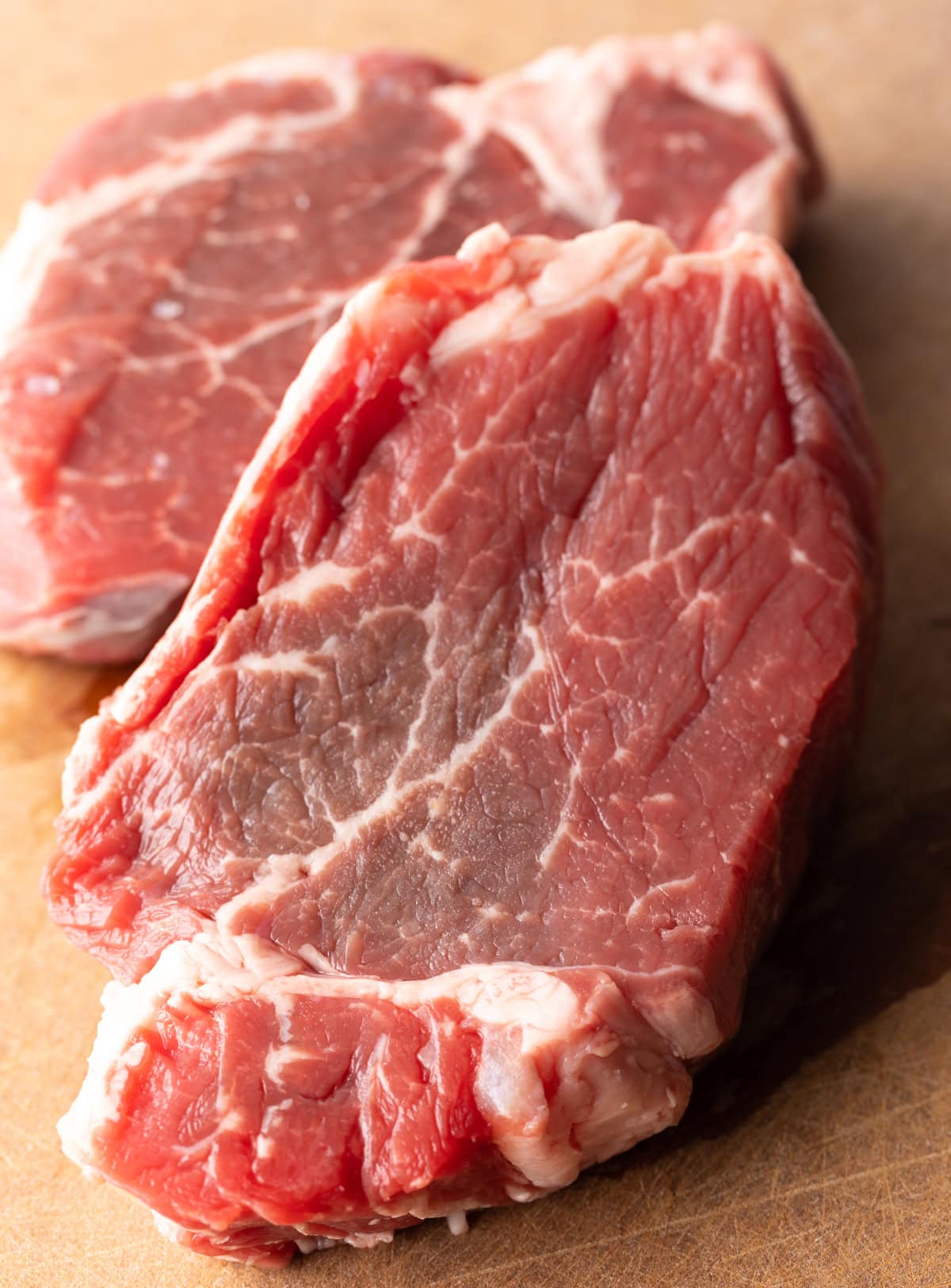 Close up of a pair of raw steaks on a brown cutting board.