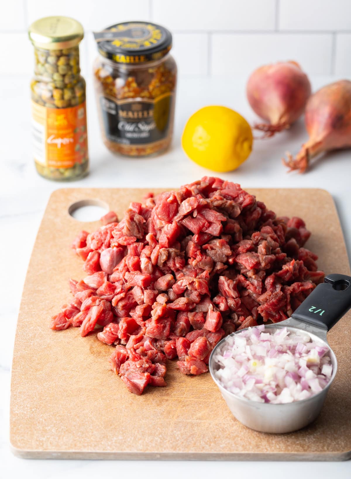 Pile of chopped steak on a wooden cutting board. There is a measuring cup of chopped shallot in front of it, and the rest of the ingredients needed to make steak tartare in the background.