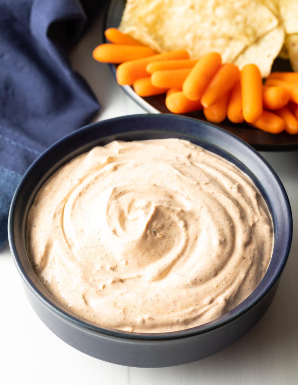 blue bowl with creamy taco dip, plate of carrots and chips in the background