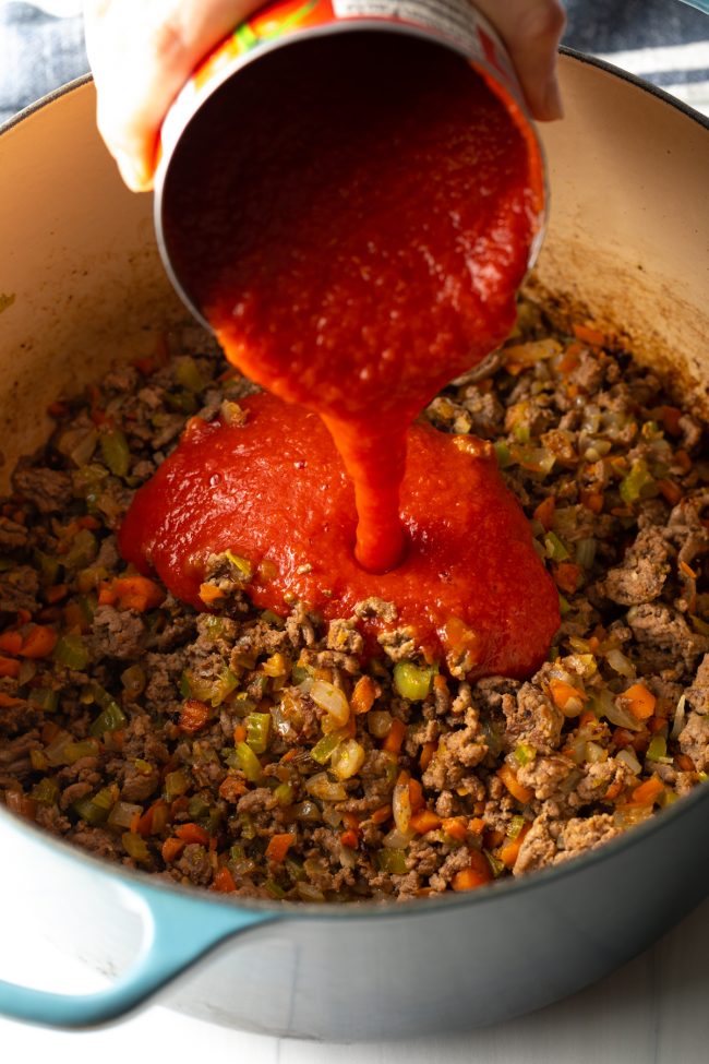 pouring canned tomato sauce to the large pot of cooked ground beef and veggies