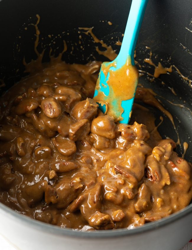 stirring pecan halves with blue spatula into homemade caramel in a large pot