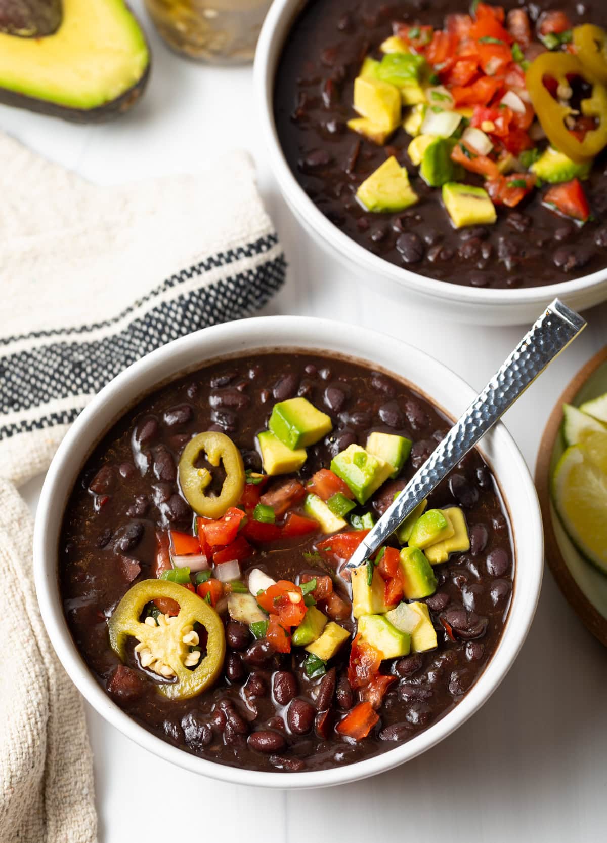 White bowl of black bean soup topped with slices of jalapenos, chopped avocado, and pico de gallo.
