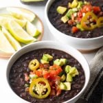 Two white bowls of black bean soup topped with slices of jalapenos, chopped avocado, and pico de gallo.