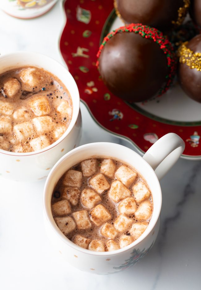 two mugs of hot cocoa in christmas mugs with plate of chocolate balls on festive plate in the background