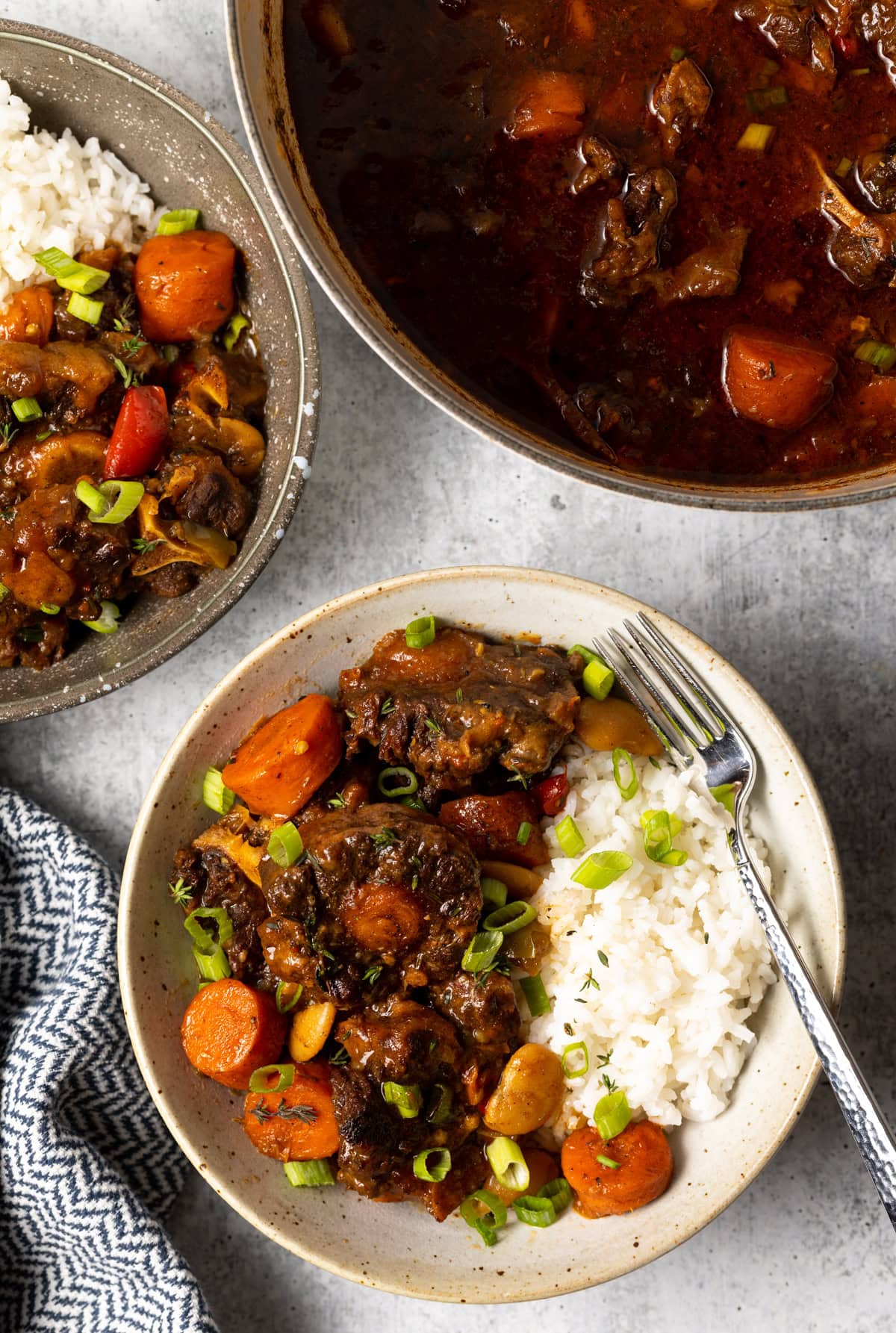 Top view two bowls with homemade oxtail stew and chopped scallions, white rice and soup pot in background.