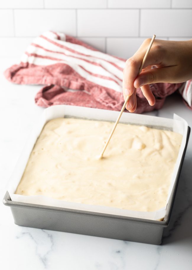 raw cake batter in metal square tin, using a skewer to remove bubbles