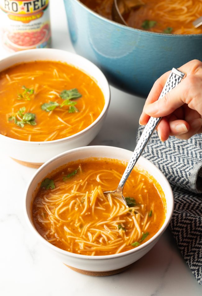 two bowls of sopa de fideo with pot of soup in background, foreground has hand holding spoon and scooping up a bite of soup
