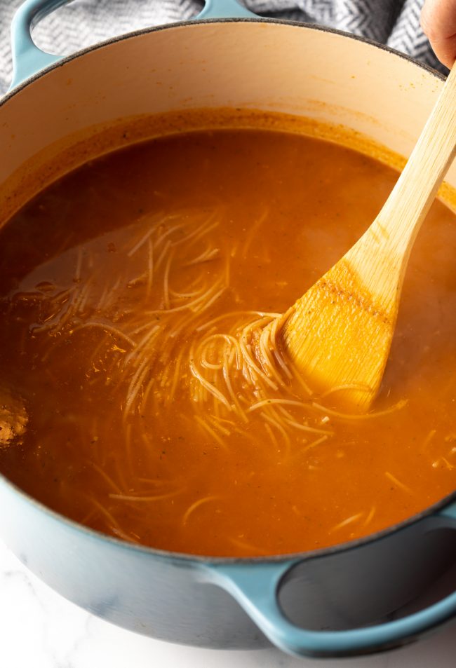 wooden spoon stirring Mexican tomato soup with noodles in a large pot