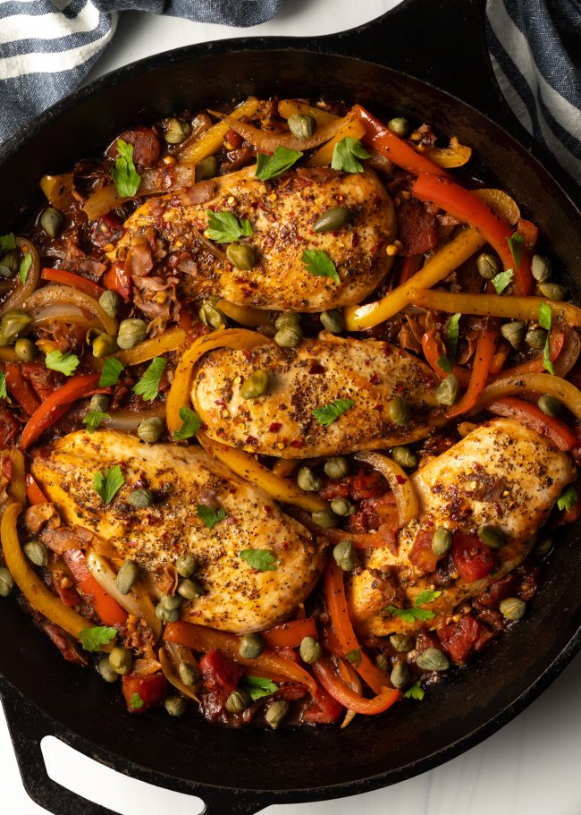 top view chicken breasts in a cast iron skillet with peppers, onions, capers, and prosciutto 