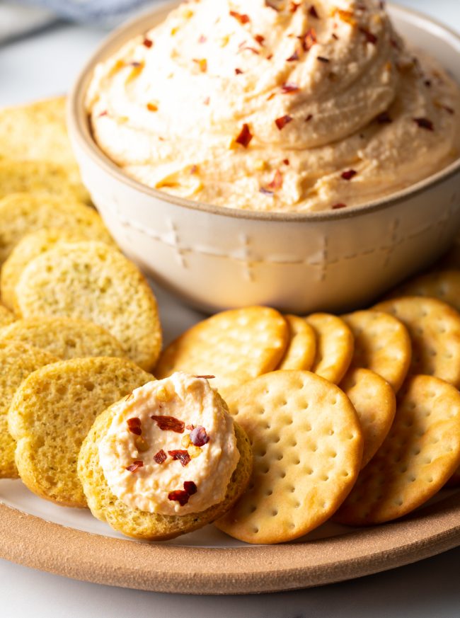 close view platter with crackers and bread slices, bit of cheese spread on one cracker with full bowl in the background