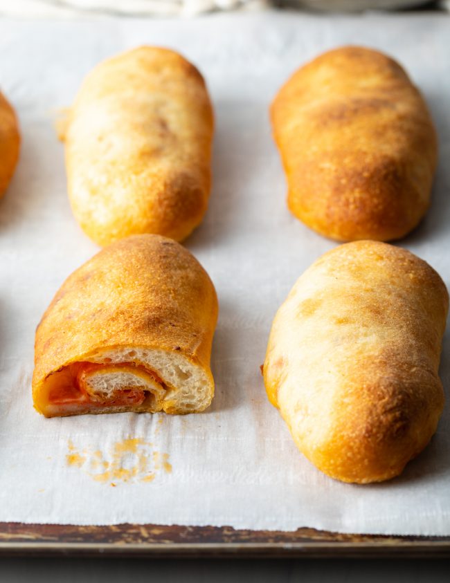 side view of sliced open pepperoni roll on a baking sheet with whole loaves