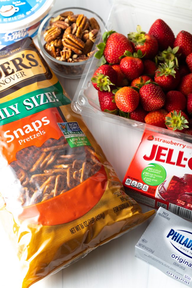 top view basket of fresh strawberries, box of strawberry jello, box of cream cheese, bag of pretzels, bowl of pecans, tub of cool whip