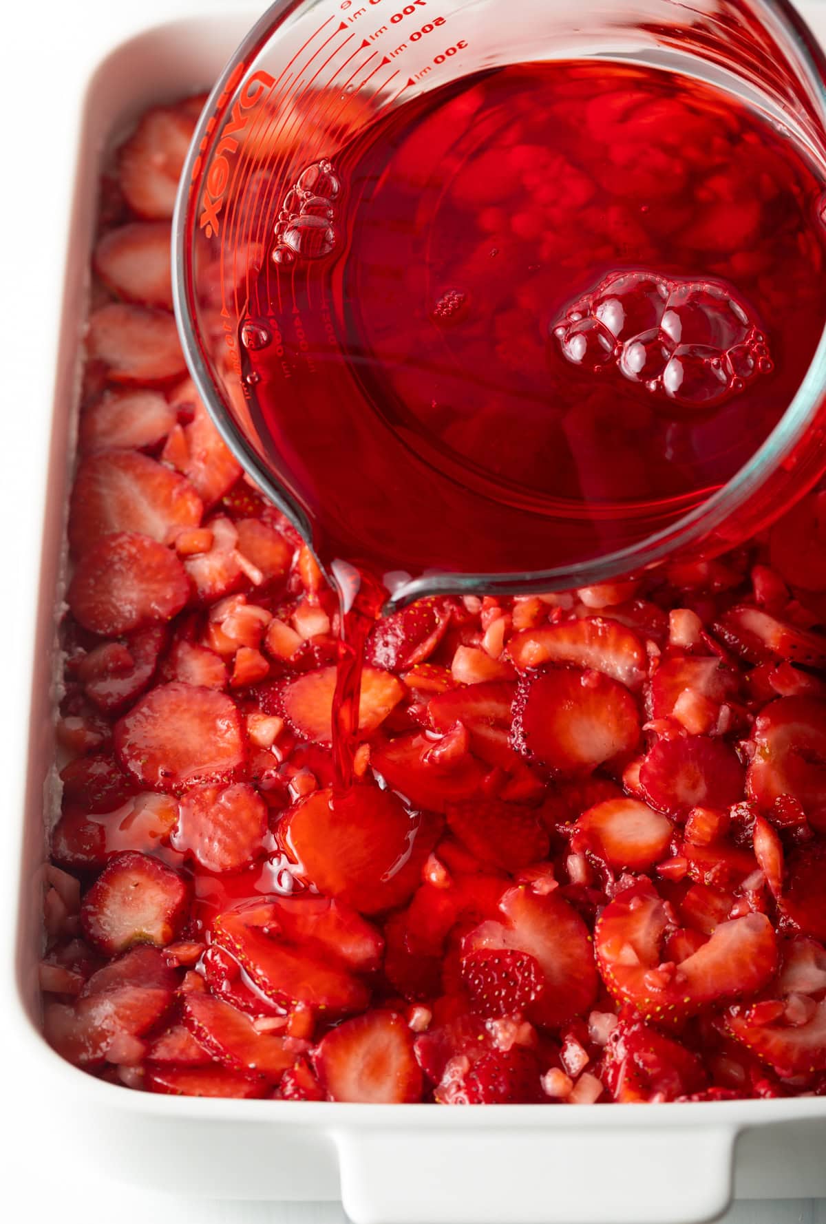 Pouring the red jello mixture over the layers of diced strawberries in the strawberry pretzel dessert recipe.