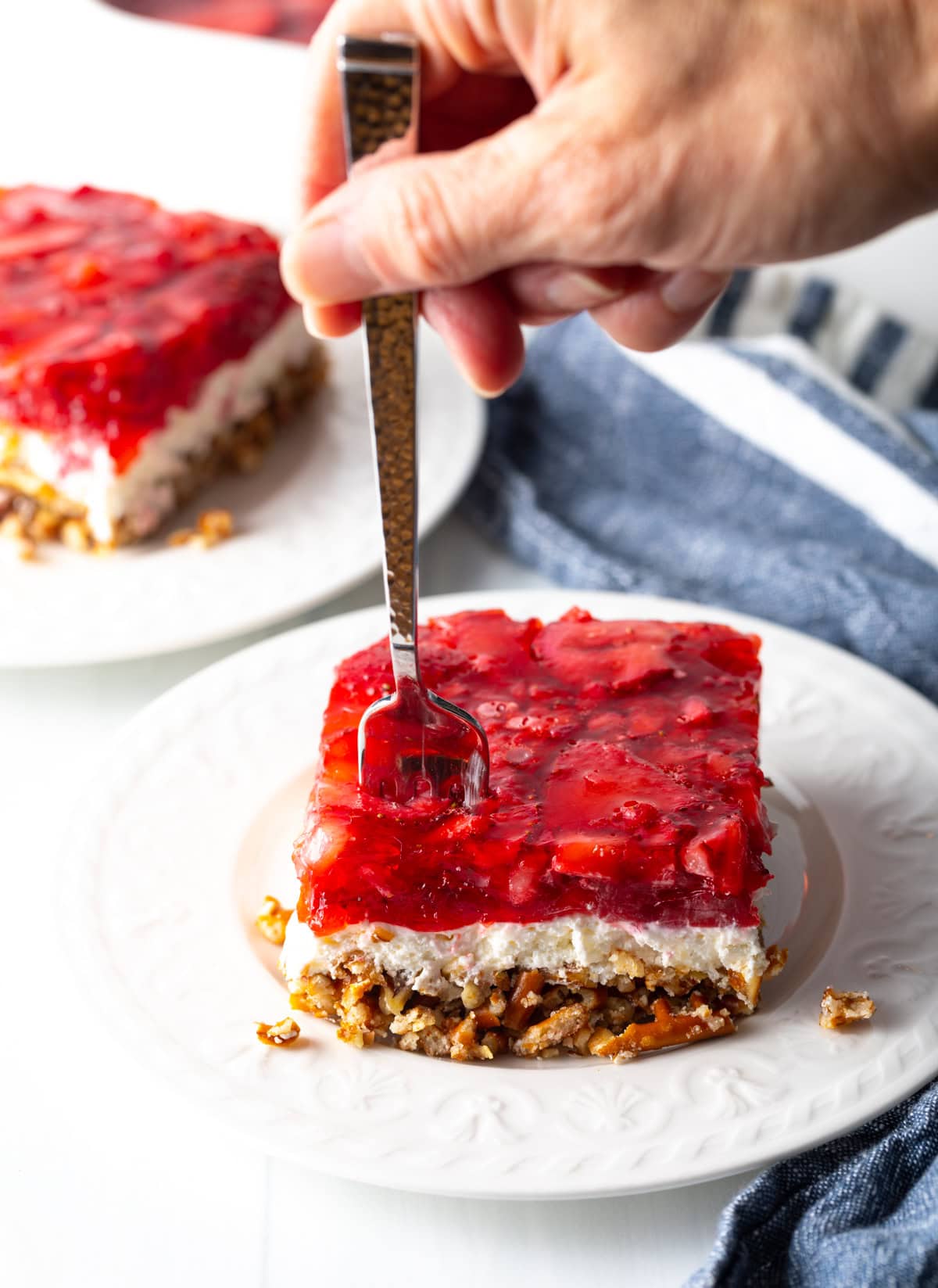 Hand with fork actively taking a bite of the layered strawberry jello cream cheese fluffy and pretzel pecan crust dessert.
