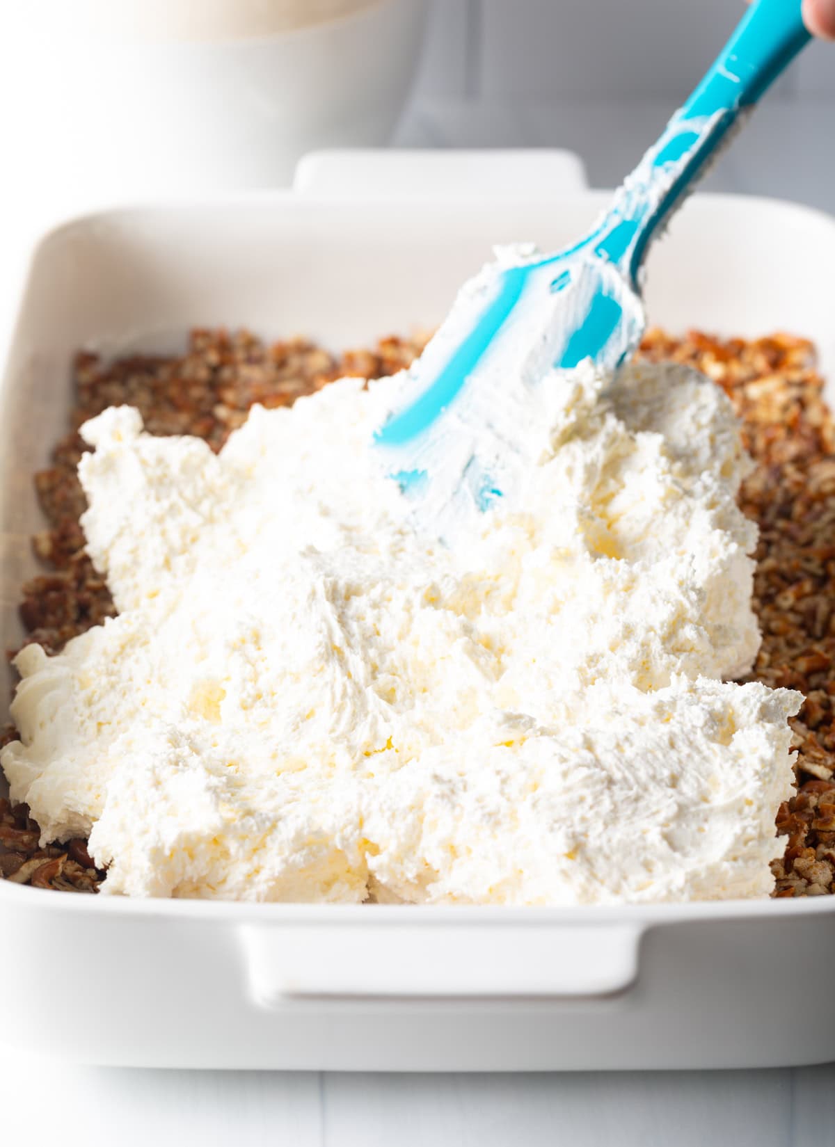 Using a blue spatula to spread cream cheese cool whip fluff across the pretzel crust.