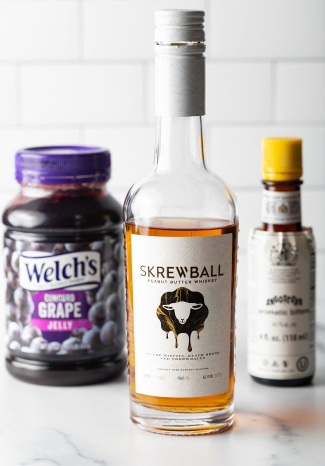 Bottle of Skrewball whiskey, jar of grape jelly, and bottle of bitters.