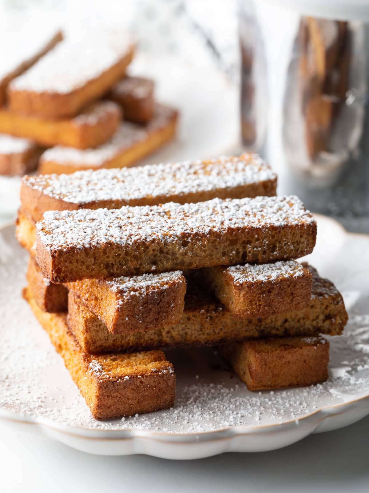 Sticks of air fryer French toast are stacked Lincoln-log-style on a white plate, topped with powdered sugar.
