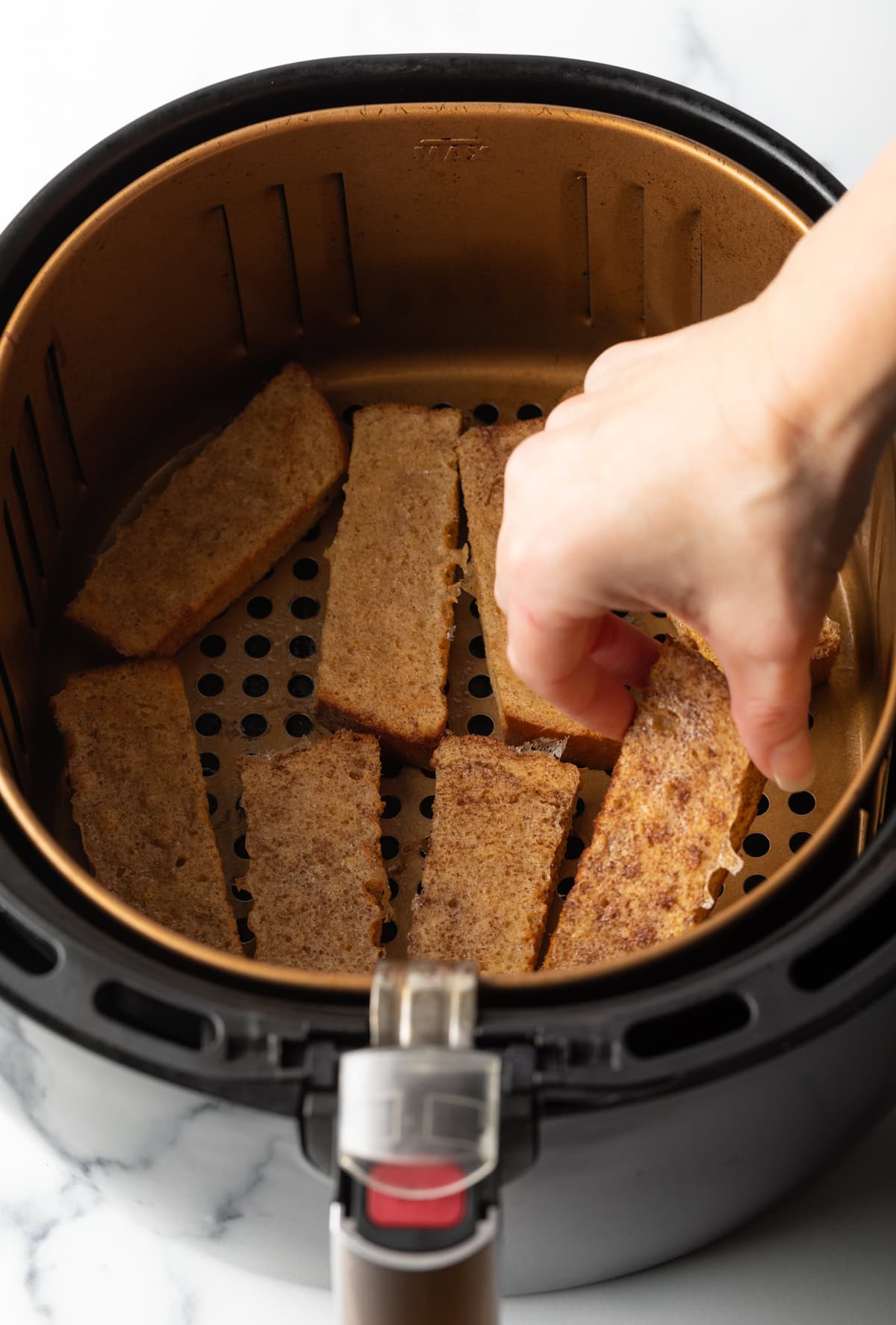 A hand is placing French toast sticks in an air fryer basket in a single layer.