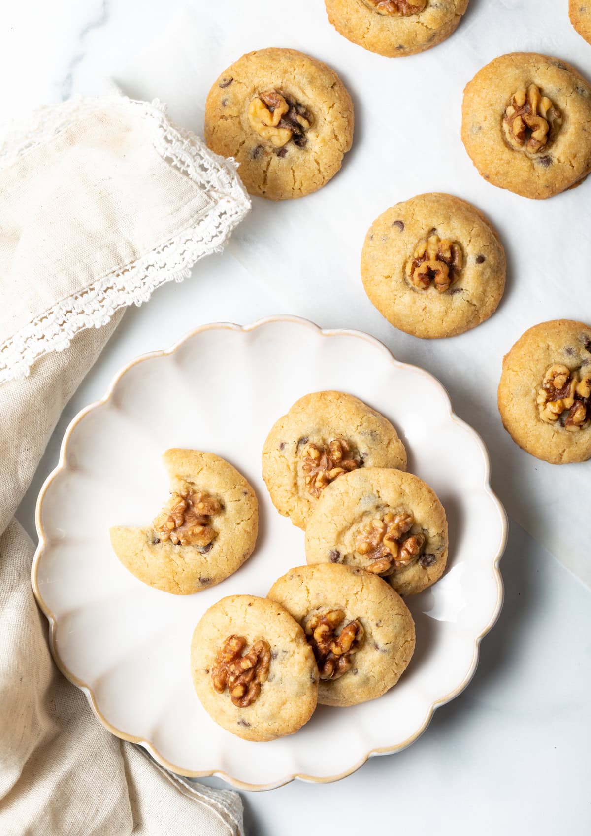 Overhead shot of chocolate chip and walnut cookies stacked in a semi-circle on a white plate, with one cookie that has a bite taken out of it.