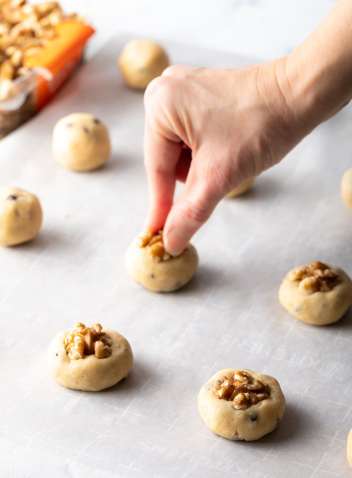 A hand is pressing a halved walnut into the balls of chocolate chip and walnut cookie dough.