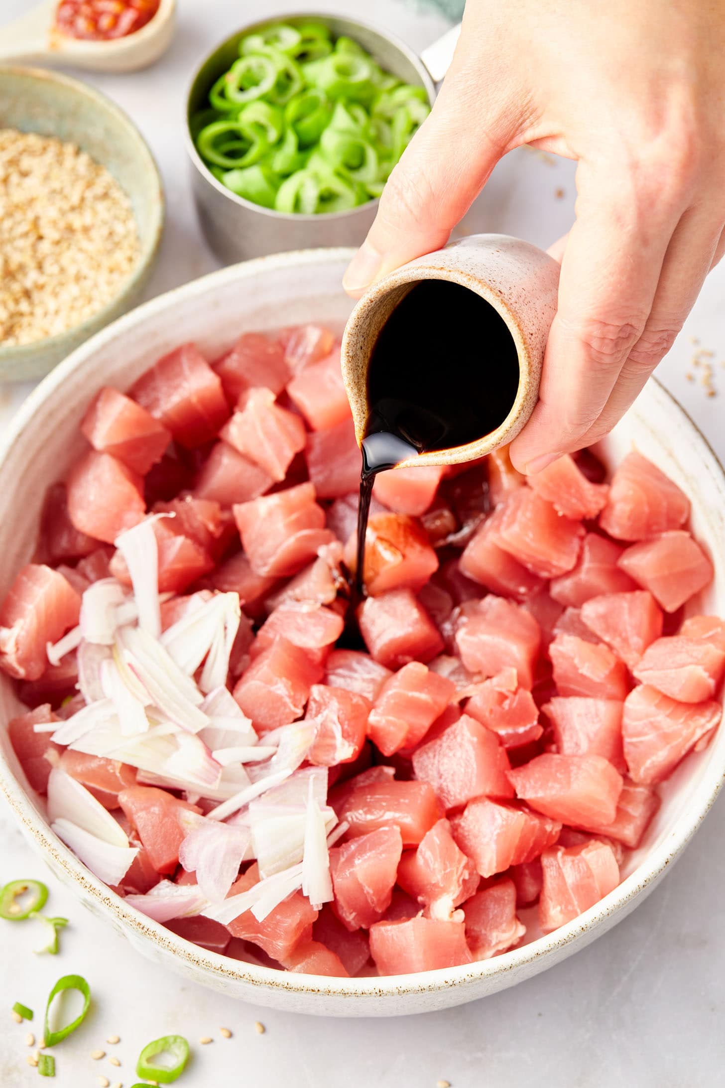 A hand pouring a small pourer of soy sauce into a bowl of diced tuna and sliced onions.