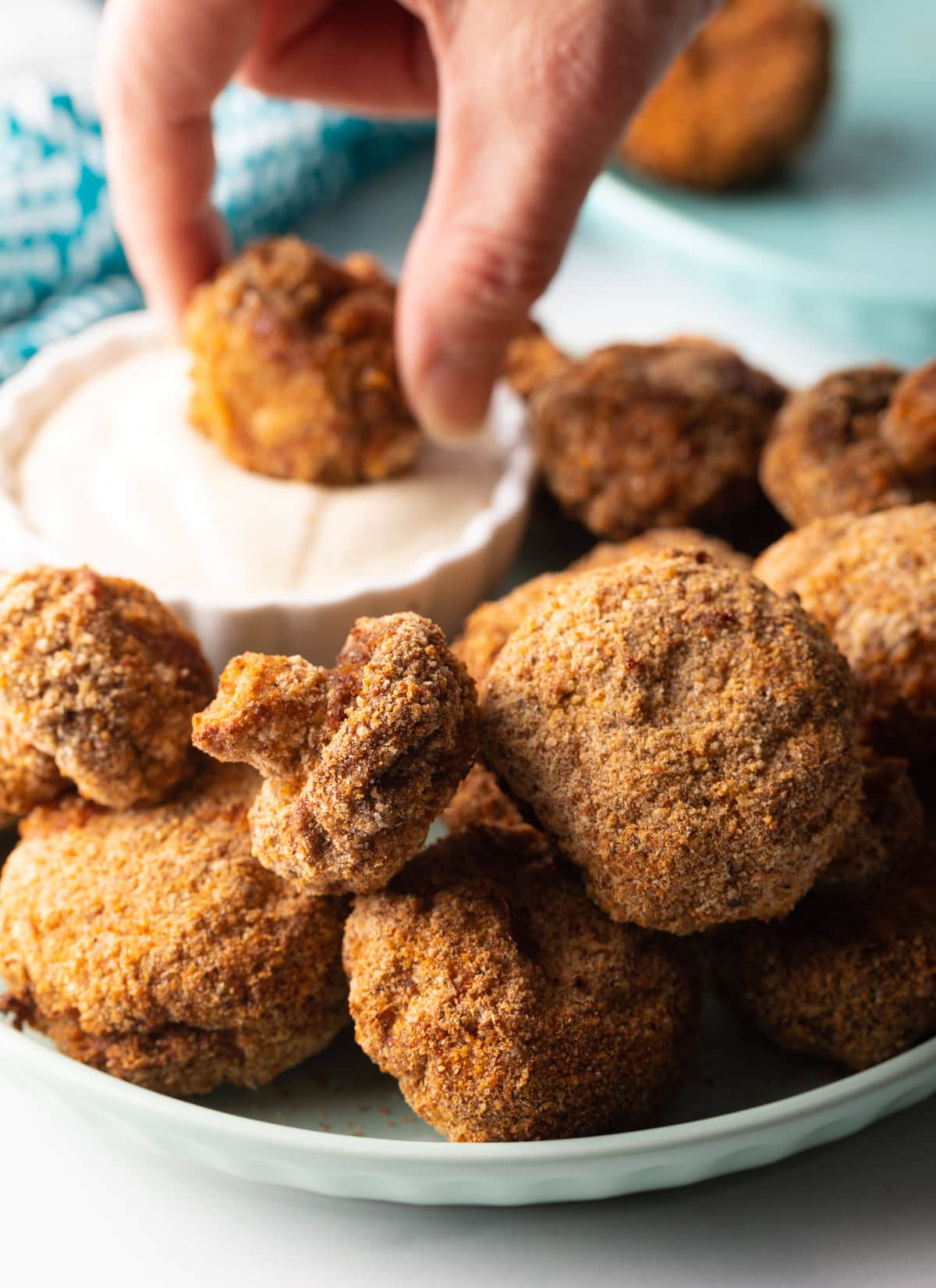 Close view of breaded air fried mushrooms on a plate. In the background is a hand dipping a mushroom into a white sauce.