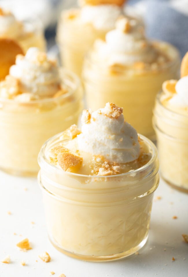 Closeup of vanilla pudding in a glass jar topped with cream and crumbled biscuits.