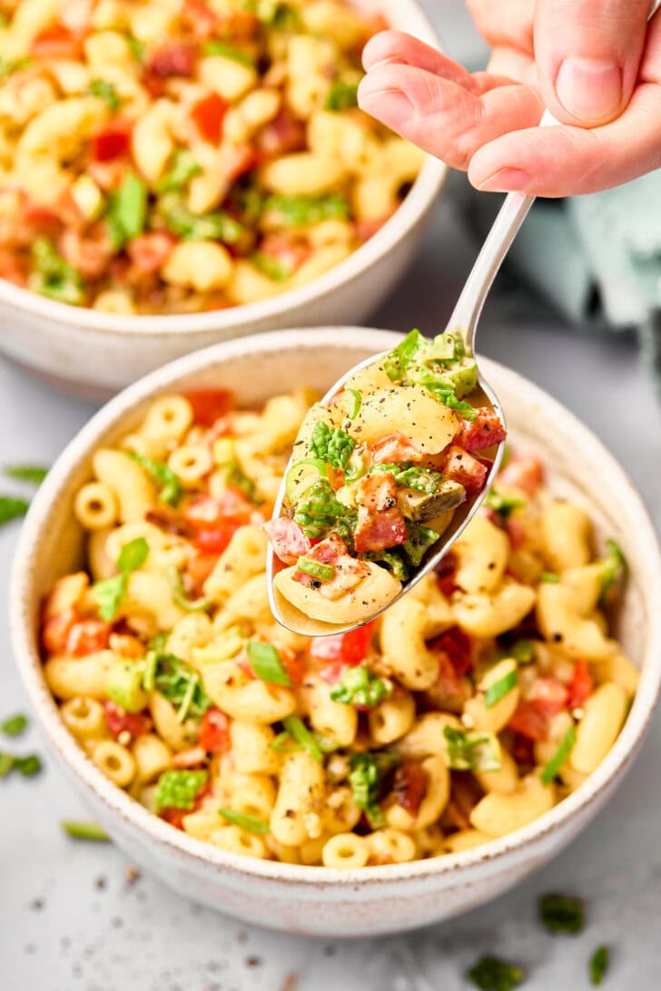 A spoon held above two bowls of macaroni salad to show chunks of tomato, roasted pepper, sweet pickle, and chopped kale.