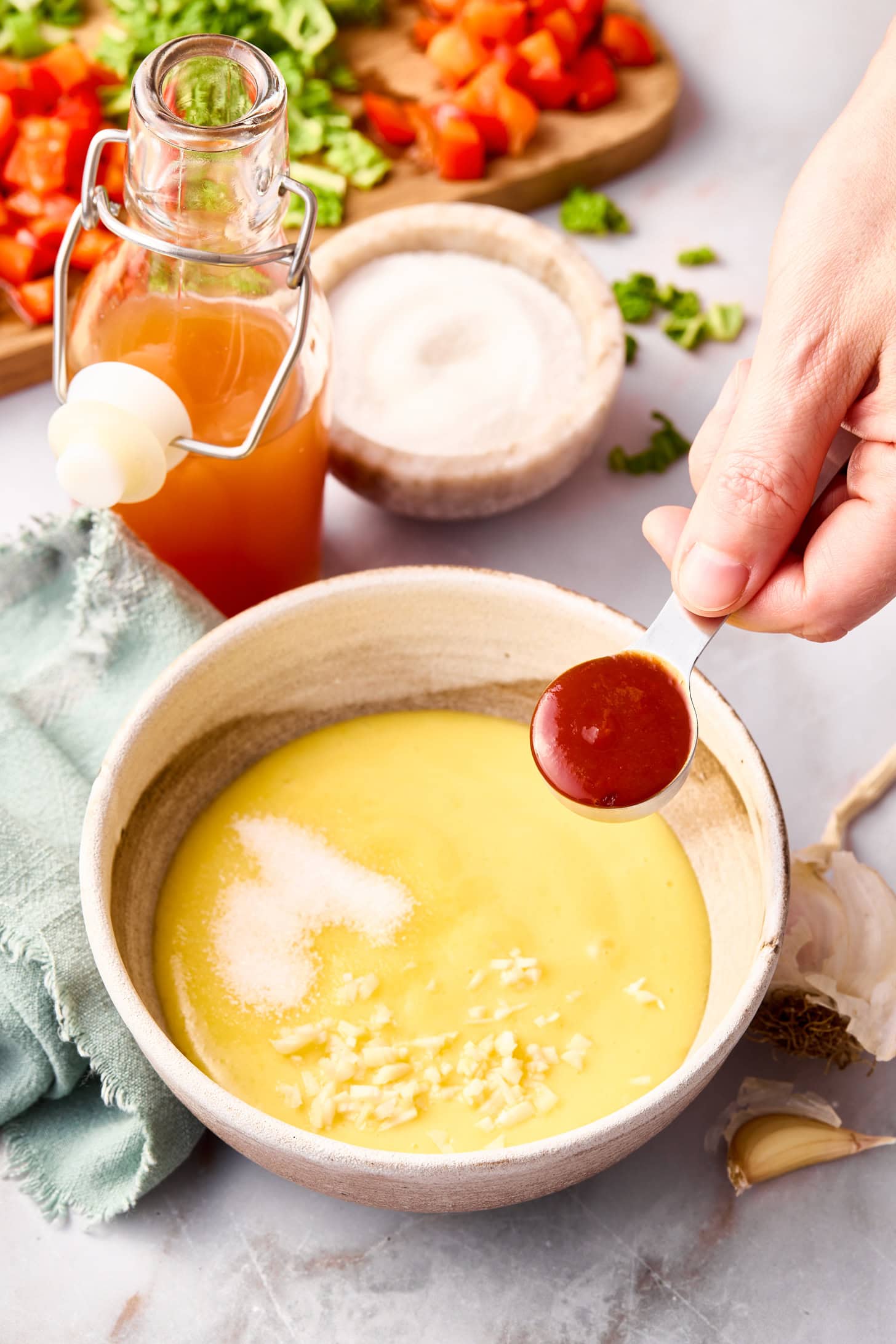 A hand holding a spoon of hot sauce over a bowl of mayonnaise dressing. Other ingredients for a pasta salad and a bottle of apple cider vinegar are visible in the background.