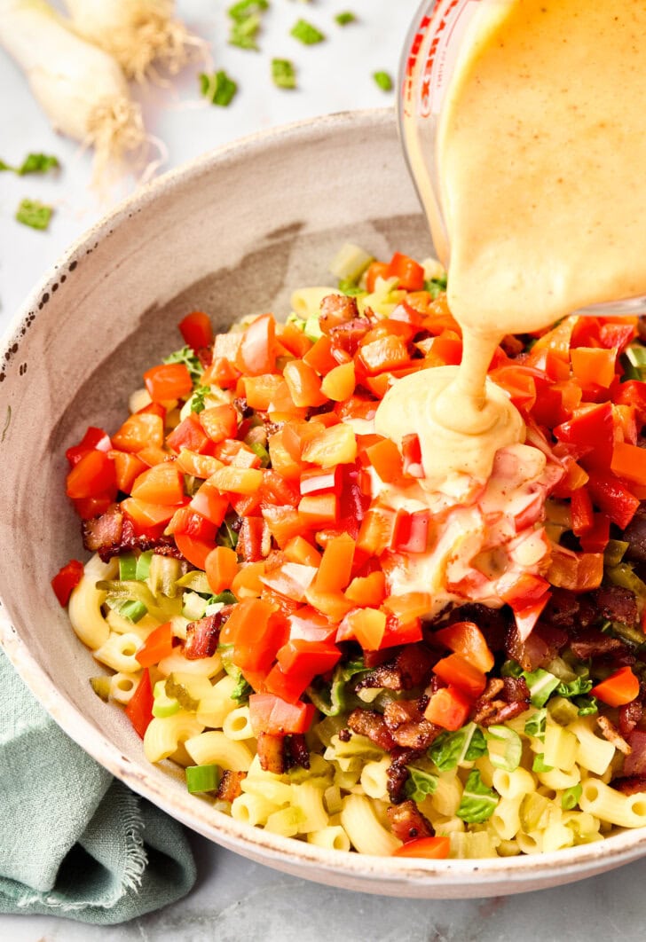 Mayonnaise-based dressing being poured from a pyrex jug into a bowl of chopped ingredients for macaroni salad.