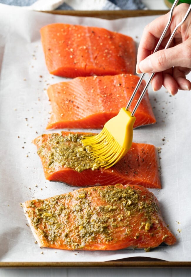 Hand with basting brush, brushing pesto over the tops of 4 raw salmon fillets.