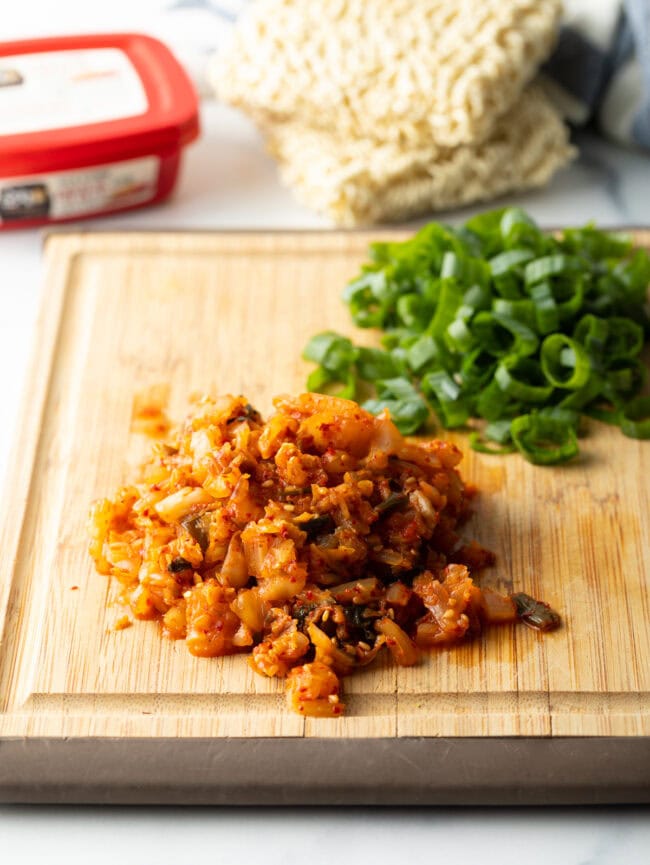 Wooden cutting board with pile of kimchi and pile of sliced green onions.