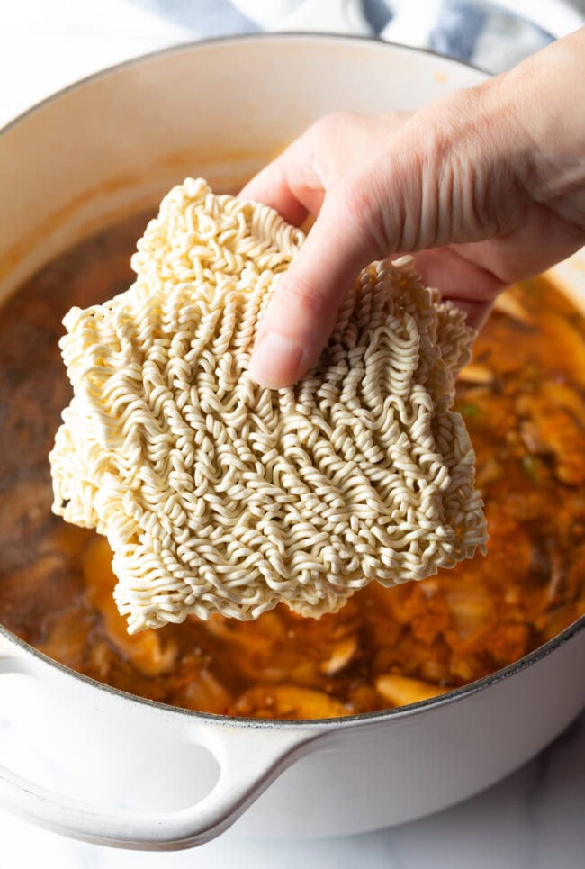 Hand adding two packets of raw ramen noodles to the cooking pot.