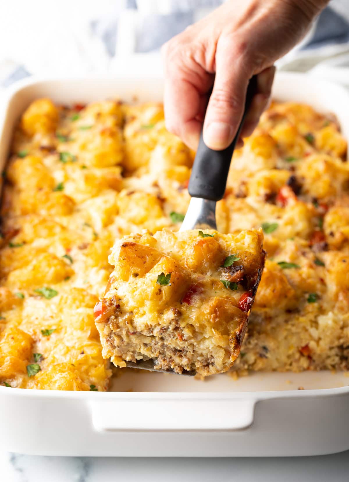Hand holding spatula as it lifts a wedge of tater tot breakfast casserole from the dish.