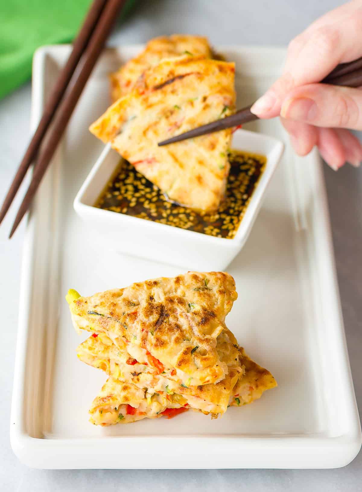 Korean Pancake Recipe #ASpicyPerspective #KoreanPancakes #KoreanPancakeRecipe #Pajun #Pajeon #Korean #Vegetarian A hand is dipping a pancake made from veggies into sauce.