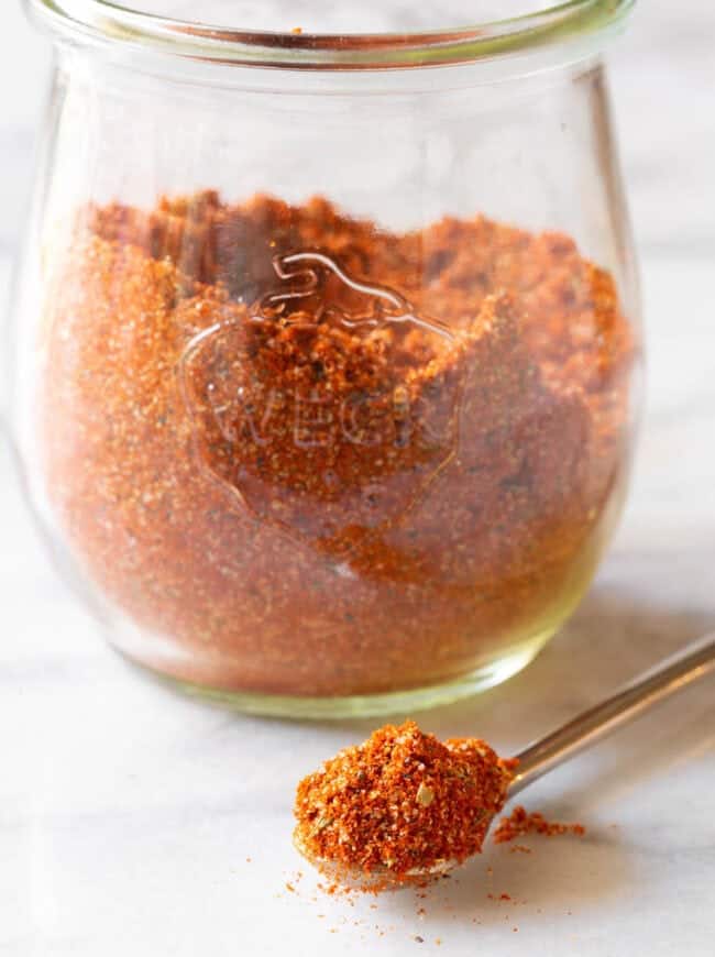 A glass jar full of blackened seasoning and in front of it a metal spoon with a spoonful of blackened seasoning. 

