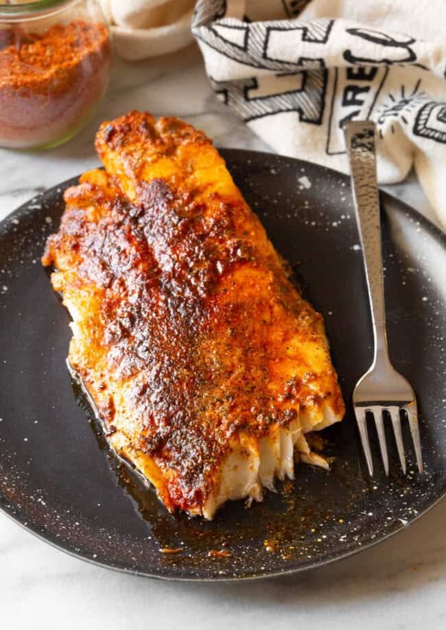 A cooked filet of fish covered in blackened seasoning, on a black plate with a metal fork. 