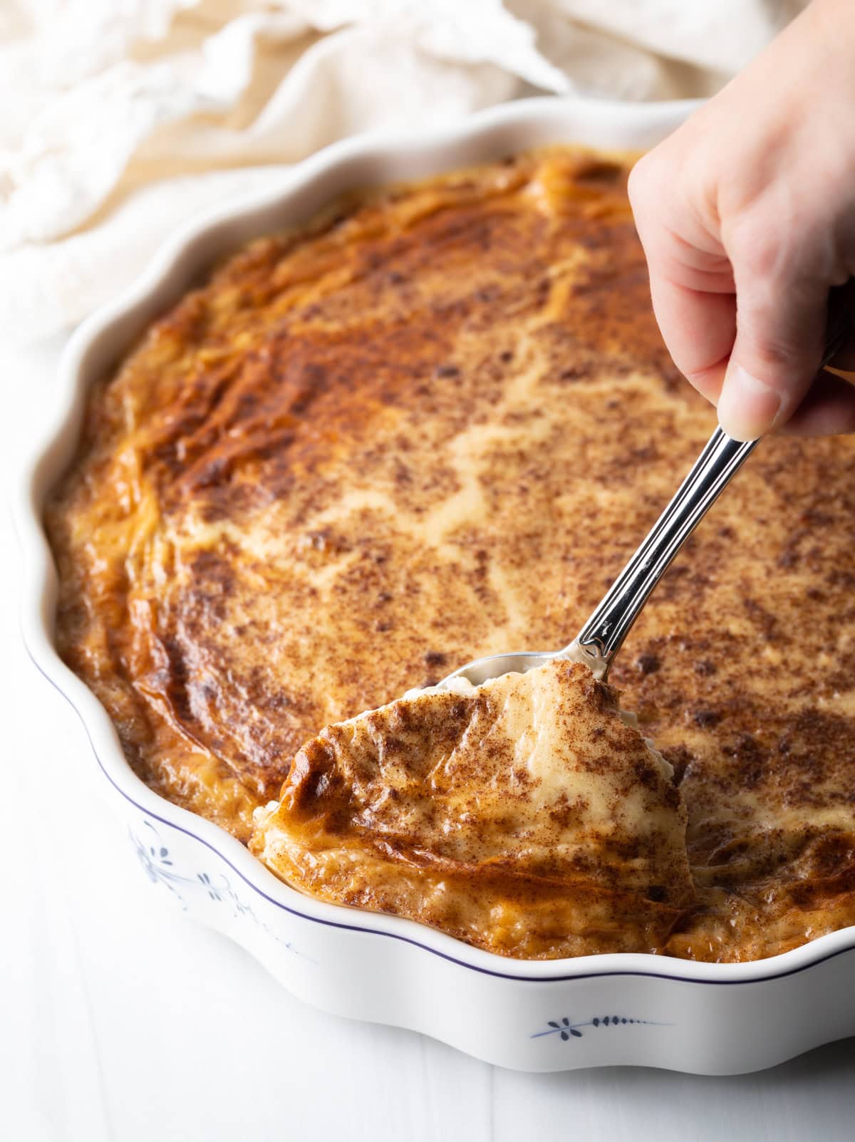 Hand with metal spoon scooping a serving of rice pudding from a large baking dish.