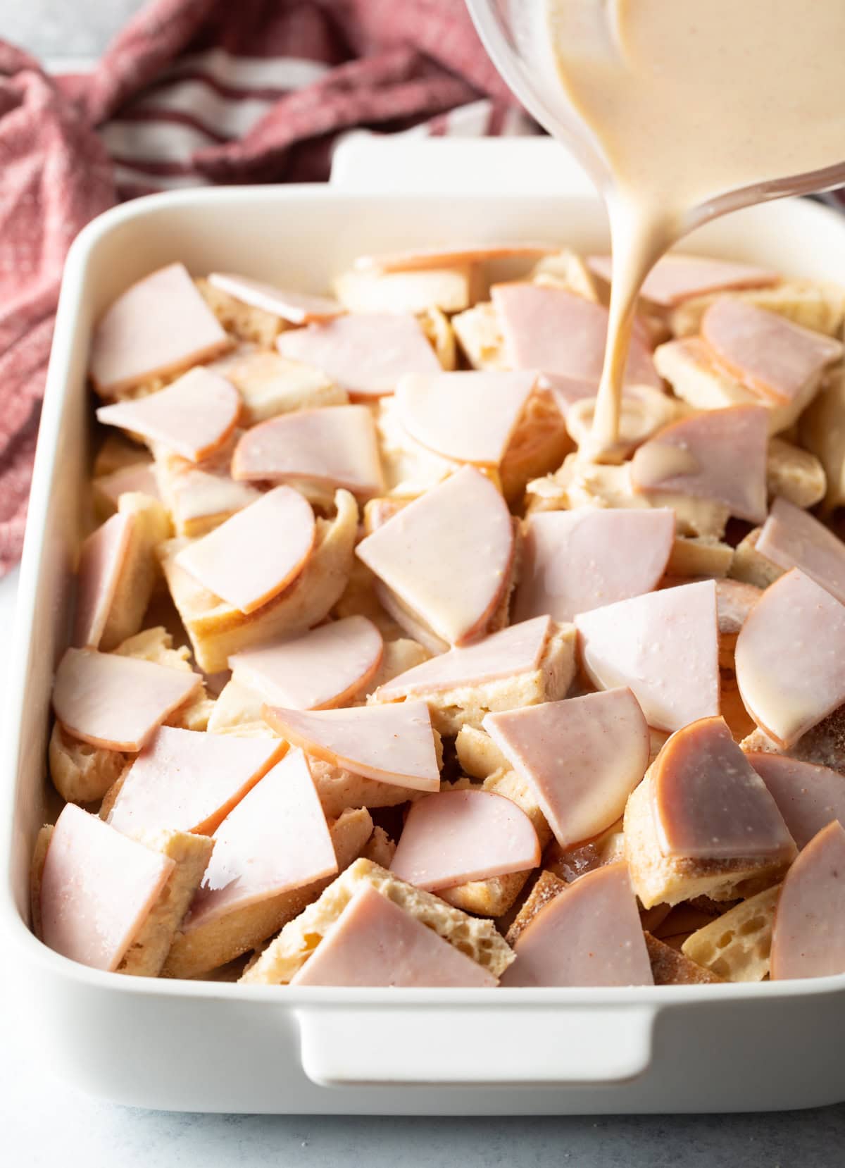 A white baking dish with quartered English muffins and Canadian bacon with egg mixture being poured on top.
