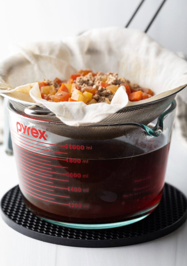 A strainer holding the consomme raft over a liquid measuring cup.