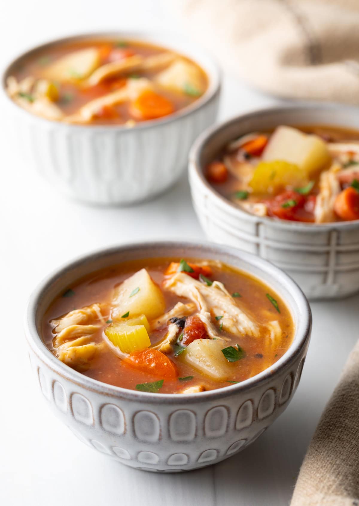 3 small white bowls of chicken stew with potatoes, carrots, and celery in a tomato broth.