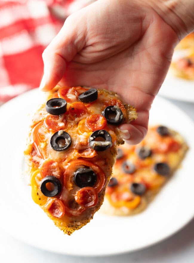 Hand holding chicken cutlet topped with pizza toppings.