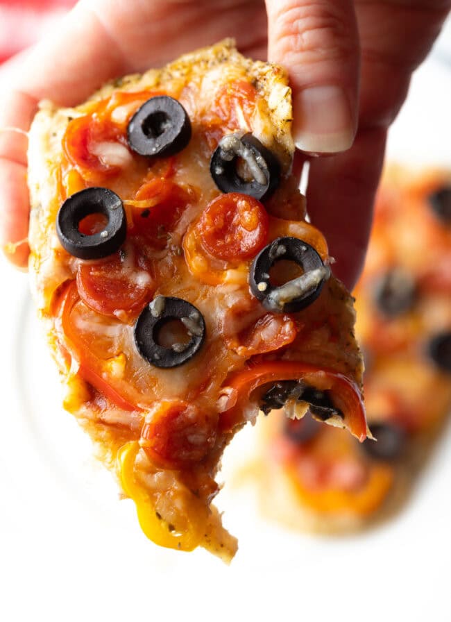 Hand holding keto chicken crust pizza piece to camera, with bite taken out.