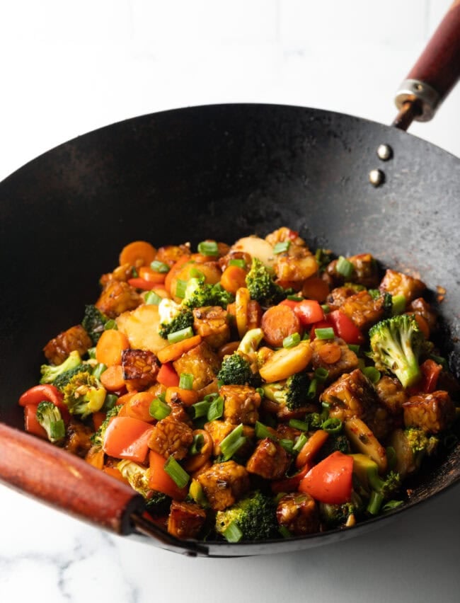Sauteed tempeh cubes, chopped veggies, and stir fry sauce cooking in a wok.