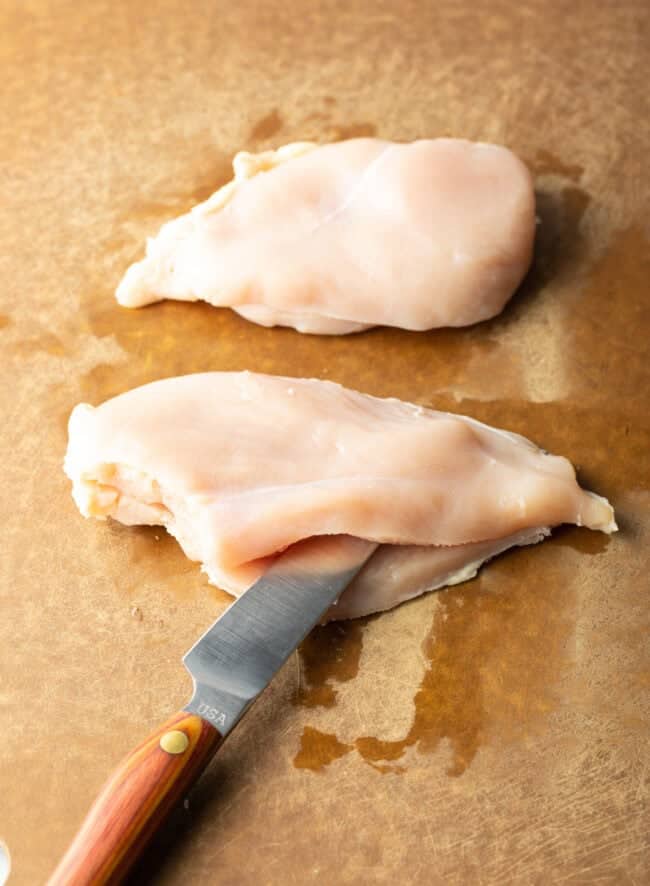 Large knife slicing raw chicken breast in half width-wise.
