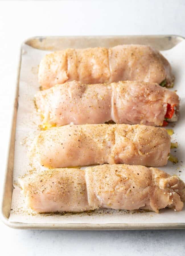 Four stuffed and rolled chicken breasts on a baking tray. They have been seasoned with olive oil and seasonings.
