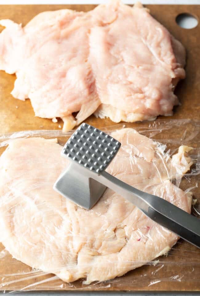 Butterflied chicken breasts on a cutting board, covered in plastic and with a metal mallet pounding them thin.
